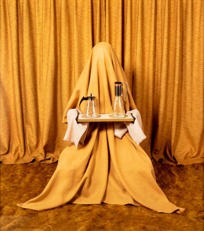 Photograph of a figure covered in yellow cloth, holding a tray with two glass pitchers